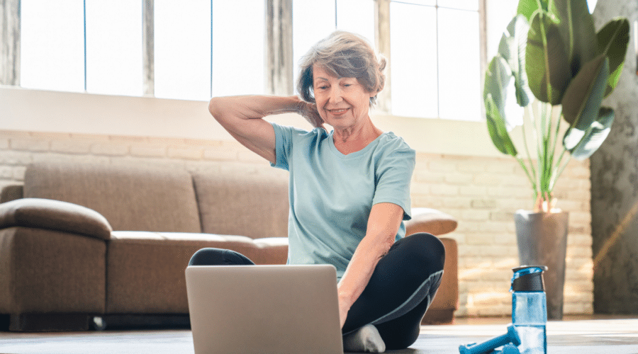 Sporty grandmother doing sport on fitness mat in training outfit while using laptop for online video